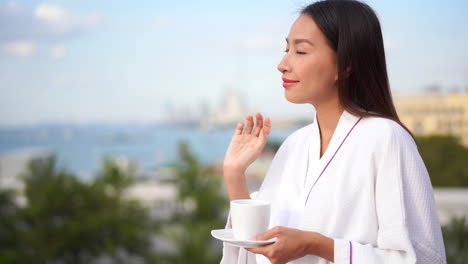 Pretty-young-Asian-woman-in-white-cotton-robe-with-ceramic-cup-in-hand-stands-on-balcony-looking-out-over-island-in-morning-light