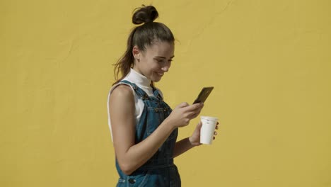 Happy-Young-Latin-Woman-holding-a-coffee-cup-takes-a-selfie-with-the-mobile-wearing-an-Overalls-with-a-yellow-background-wide-angle-shot