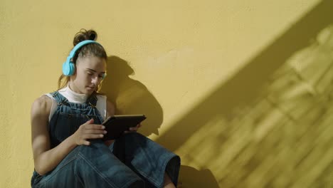Young-attractive-Woman-using-her-Ipad-while-listening-to-music-with-wireless-headphones-wearing-Overalls-seated-against-a-yellow-wall-on-a-sunny-day-medium-shot