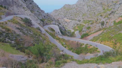 A-slow-mo-long-view-of-cyclist-moving-on-the-zigzag-road-at-mountains-with-lush-forest-in-winters-at-Mallorca,-Spain
