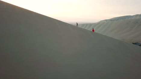 Aerial,-tracking,-drone-shot,-of-a-couple-walking-on-a-sand-dune,-revealing-the-Huacachina-oasis-village,-on-a-sunny-evening,-in-southwestern-Peru