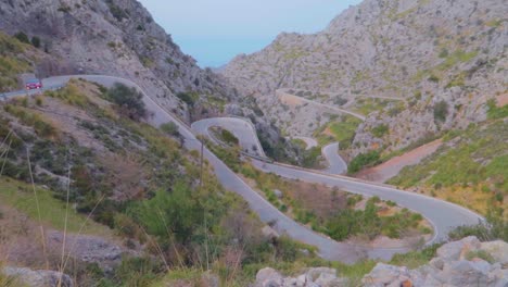 A-huge-long-view-of-a-zigzag-road-with-mountains-series-with-lush-forest-in-winters-where-a-red-car-is-moving-gently-at-Mallorca,-Spain
