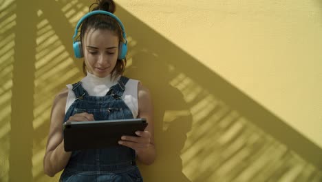 Young-brunette-Woman-using-her-Ipad-while-listening-to-music-with-wireless-headphones-wearing-Overalls-seated-against-a-yellow-wall-on-a-sunny-day-medium-shot