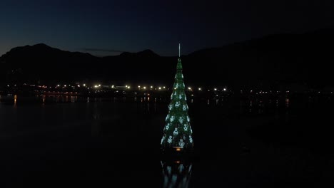 Schwimmender-Weihnachtsbaum-Mit-Lichtern-In-Rio-De-Janeiro-Bei-Sonnenuntergang-Mit-Der-Stadtbildsilhouette-Dahinter-Und-Reflexion-Des-Baums-Im-Stadtsee