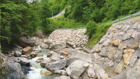 Shallow-clear-river-with-stones-surrounded-by-rocks-and-greenery-by-the-traffic-road