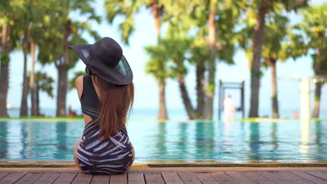 A-woman-with-her-back-to-the-camera,-in-a-bathing-suit-and-floppy-sun-hat,-sits-on-the-edge-of-a-pool-looking-out-at-the-ocean