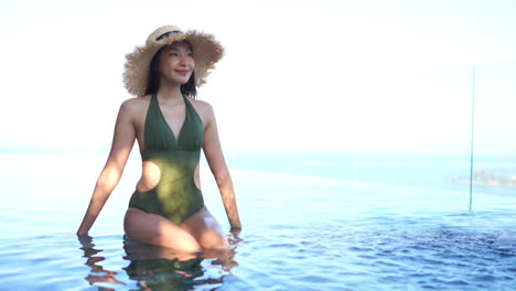 Young-attractive-Asian-woman-in-green-one-piece-bathing-suit-and-wide-straw-hat-sitting-on-edge-of-pool
