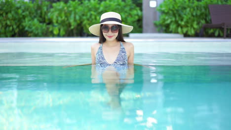 Woman-wearing-hat-and-sunglasses-relaxing-in-a-warm-clear-swimming-pool-with-tropical-landscaping-in-background
