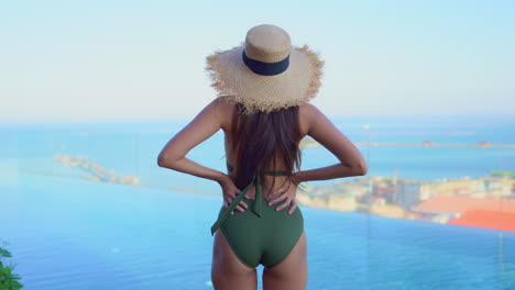 Back-view-of-a-female-tourist-standing-near-a-swimming-pool-with-her-hands-on-her-hips,-wearing-a-sun-hat-and-olive-swimsuit-,-looking-at-the-ocean-in-the-background