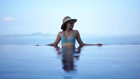 Beautiful,-elegant-woman-in-fashionable-swimwear-sitting-in-infinity-pool,-blurred-background,-soft-light
