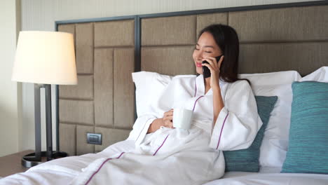 wide-shot-of-young-adult-Asian-woman-in-white-bathrobe-on-a-hotel-double-bed-holding-a-cup-of-tea-coffee-and-having-a-joyful-happy-conversation-on-her-mobile-phone