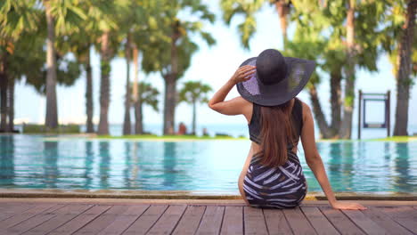 Beautiful-long-haired-woman-sits-at-the-edge-of-pool-wearing-swimsuit-and-hat