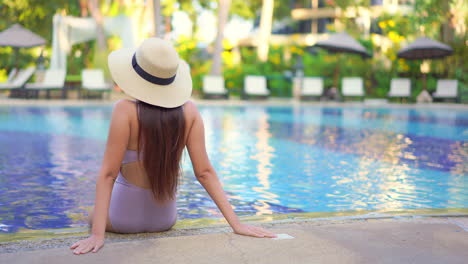 A-woman,-in-a-bathing-suit-and-Panama-hat,-with-her-back-to-the-camera-sits-on-the-pool-edge-relaxing
