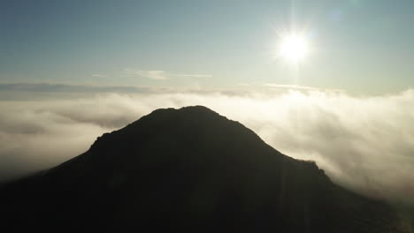 Mountaintop-peaking-high-above-fog-and-clouds-with-bright-sun,-AERIAL-SIDEWAYS