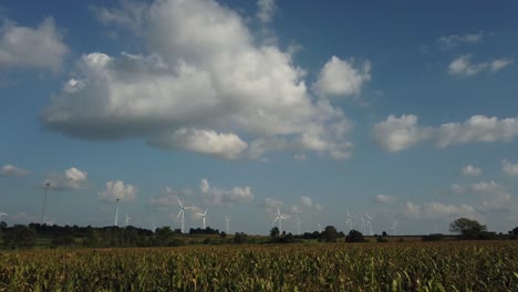 Wind-Turbines-jutting-out-of-the-horizon-of-a-beautiful-landscape,-a-cornfield-in-the-foreground-while-the-sky-is-blue-and-the-clouds-are-fluffy,-like-cotton-balls,-captured-in-slow-motion