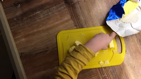 Boy-cuts-butter-with-a-knife-on-a-yellow-board,-baby-help-in-the-kitchen,-top-view