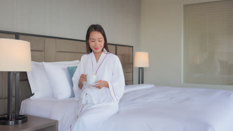 A-young-attractive-Asian-woman,-dressed-in-a-resort-bathrobe,-sits-on-the-edge-of-her-bed-as-she-enjoys-a-cup-of-coffee