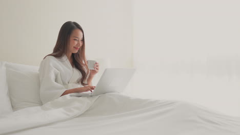 wide-shot-of-young-adult-Asian-woman-in-white-bathrobe-sitting-upright-on-a-hotel-double-bed-with-a-cup-working-on-her-laptop-joyful-happy-smiling-in-a-white-room