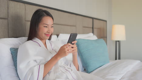 Young-adult-Asian-woman-in-white-bathrobe-on-a-hotel-double-bed-happy-texting-with-both-hands-on-mobile-phone