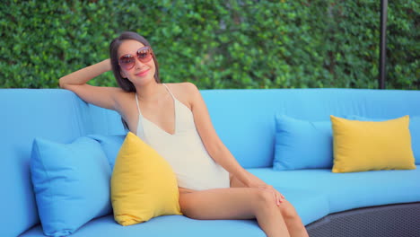 Young-asian-female-wearing-white-one-piece-swimsuit-and-sunglasses-,-sitting-on-a-big-blue-sofa-with-blue-cusions-and-complementary-yellow-cusions