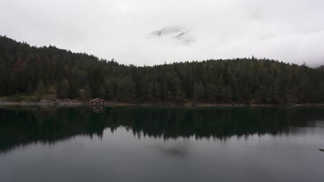 Boat-house-on-the-shore-of-a-perfect-alpine-lake,-Blindsee-with-low-clouds-partly-revealing-a-mountain-behind