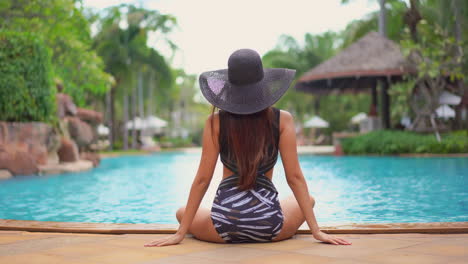 A-young-woman,-with-her-back-to-the-camera,-in-a-bathing-suit-and-floppy-sun-hat,-sits-quietly-at-the-edge-of-a-resort-swimming-pool