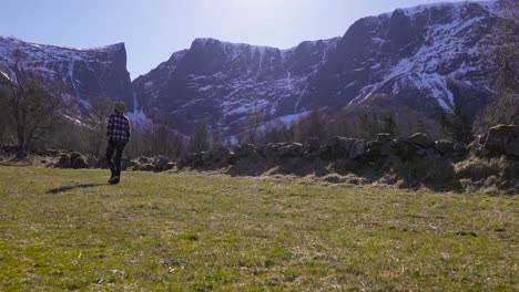 Young-man-walking-on-meadow-surrounded-by-large-snowy-mountains,-Slow-motion
