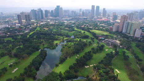 Wide-Pull-Out-Aerial-Shot-Of-Golf-Course-With-Reflection-Of-Sky-From-Pond-And-City-Skyline-At-The-Back