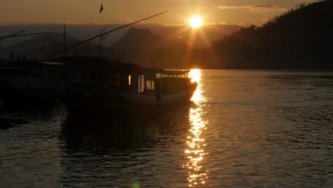 A-stunning-view-of-the-bright-orange-sunset-over-the-calm-waters-of-the-Mekong-river-in-Luang-Prabang,-Laos-with-a-docked-tourist-boat-by-the-riverbank---Wide-shot