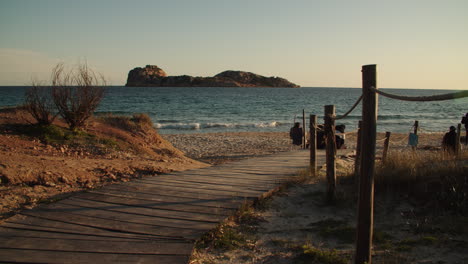 Wooden-walkway-at-beach-with-people-in-front-of-island,-sunset,-Porto-Tramatzu