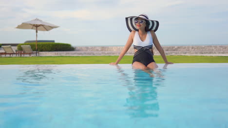 A-young-woman,-in-a-black-and-white-one-piece-bathing-suit-and-matching-floppy-sun-hat,-sits-in-the-shallow-edge-of-a-resort-pool