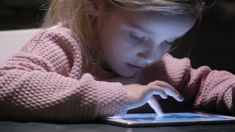Little-girl-plays-game-in-glow-of-tablet-on-table,-close-up