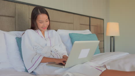 Young-Asian-woman-sitting-up-in-her-hotel-bed-and-wearing-a-robe-balances-her-laptop-on-her-legs-as-she-taps-on-the-keyboard
