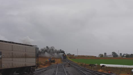 Head-on-View-of-a-Steam-Locomotive-Pulling-Freight-Pulling-into-Yard-with-Cattle-Watching-also-with-Smoke-and-Steam-on-a-Rainy-Day