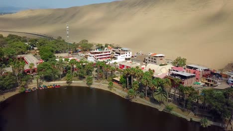 Aerial,-drone-shot,-panning-over-the-Huacachina-lagoon,-at-the-oasis-village,-on-a-sunny-day,-in-south-Peru