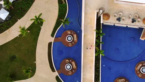 Modern-hotel-resort-with-pool,-lounges,-deckchairs-with-umbrellas,-people,-unrecognizable-walk-down-the-poolside,-luxury-vacation,-top-down-aerial-view