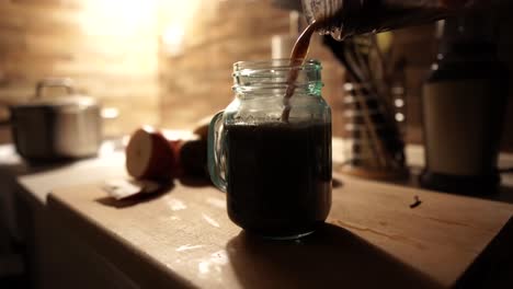Pouring-dark-cocktail-into-retro-blueish-jar-standing-on-wooden-chopping-board,-STILL,-4k