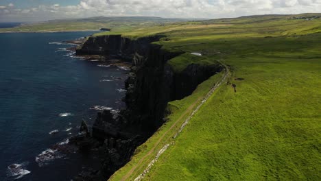 Hikers-walking-along-top-of-Cliffs-of-Moher-hiking-trail-walk,-aerial-view