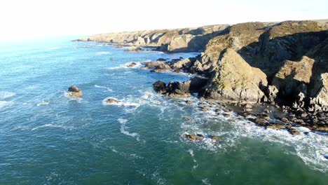 Aerial-view-following-coastal-edge-rocky-island-coastline-as-waves-crash-into-jagged-shoreline