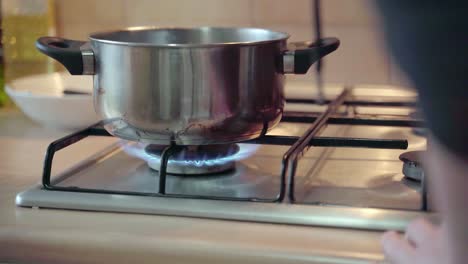 Cooking-In-The-Kitchen---Putting-On-The-Glass-Lid-Of-A-Stainless-Pot-On-The-Stove-With-Fire-On---tilt-up-slowmo-shot