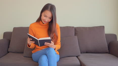Zufriedene-Asiatische-Frau,-Die-Ein-Buch-Liest,-Während-Sie-Auf-Dem-Sofa-Im-Wohnzimmer-Sitzt