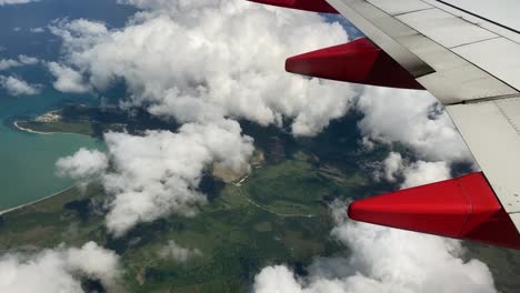 Tropical-beach-below-clouds-and-airplane-wing,-view-from-passenger-window
