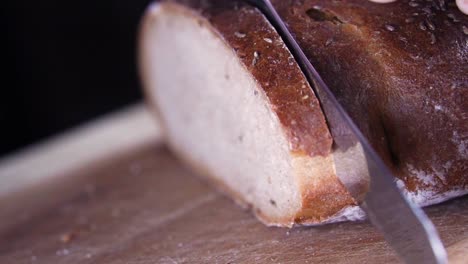 Long-serrated-blade-of-knife-slicing-through-the-crust-of-fennel-seed-bread-on-wooden-chopping-board-with-black-background,-SLOW-MOTION