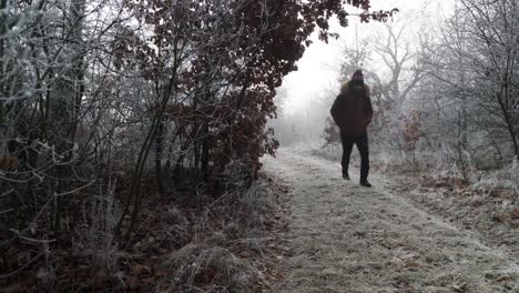 Caucasian-man-hiking-on-from-mysterious-haze-on-frost-covered-dirt-road-in-winter-forest
