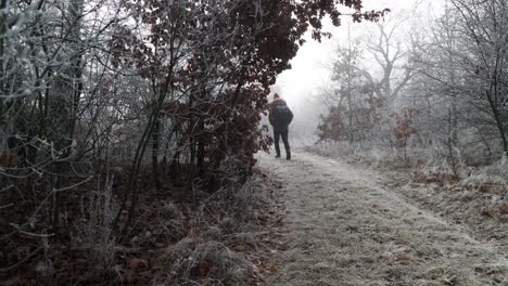 Caucasian-man-hiking-on-into-mysterious-haze-on-frost-covered-dirt-road-in-winter-forest