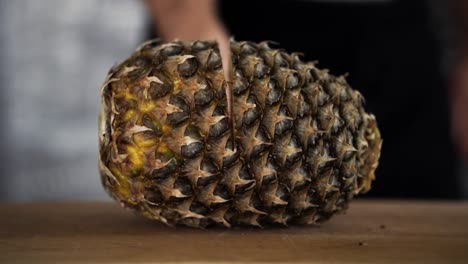 Chef-in-black-apron-cutting-into-the-middle-of-pineapple-with-big-knife-on-wooden-chopping-board,-SLOW-MOTION
