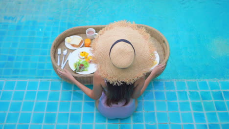 Woman-in-bathing-suit-sitting-in-swimming-pool-with-breakfast-basket