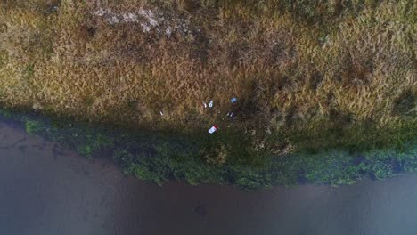 Plastic-trash-littered-on-bank-of-small-river,-overhead-rising-aerial