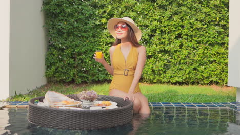 Eine-Frau-Am-Rand-Des-Pools-Hält-Einen-Schwimmenden-Teller-Fest,-Während-Sie-Ein-Glas-Orangensaft-Trinkt