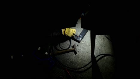 Torch-illuminating-late-night-welding-in-the-dark-using-a-handheld-tool-to-make-a-joint-for-a-metal-bed-frame-on-a-workshop-floor-with-a-hammer-nearby
